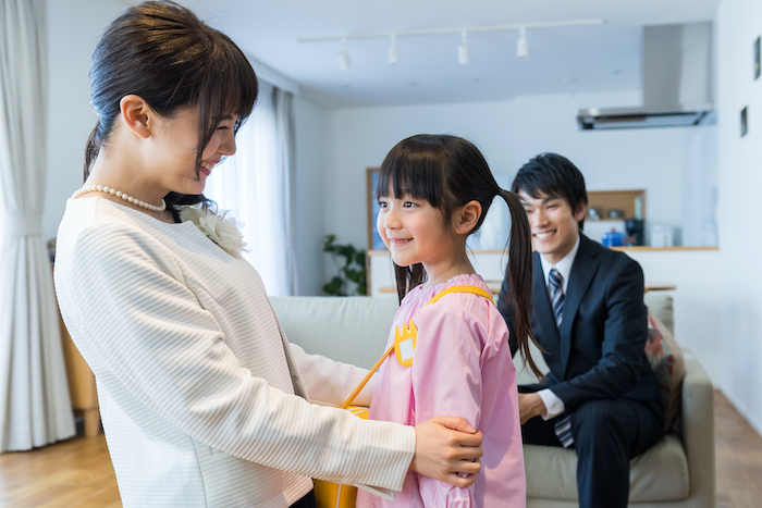 園服を着て立つ少女とそれを見てほほ笑む両親